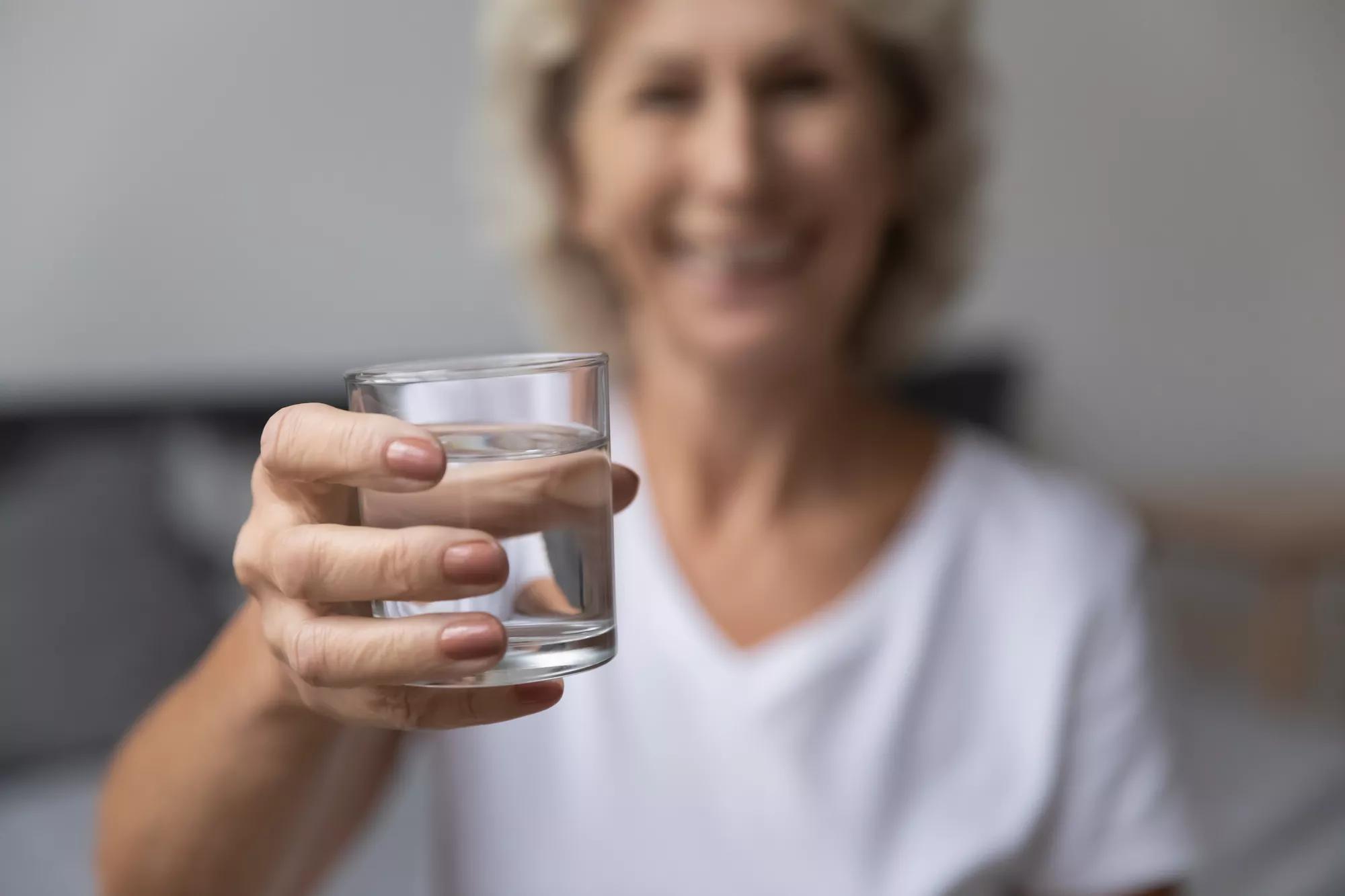 Elderly woman stretch arm to camera with glass of still or mineral water, close up focus on aqua and hand. Concept of healthy lifestyle, dehydration prevention, boost metabolism, skin and health care