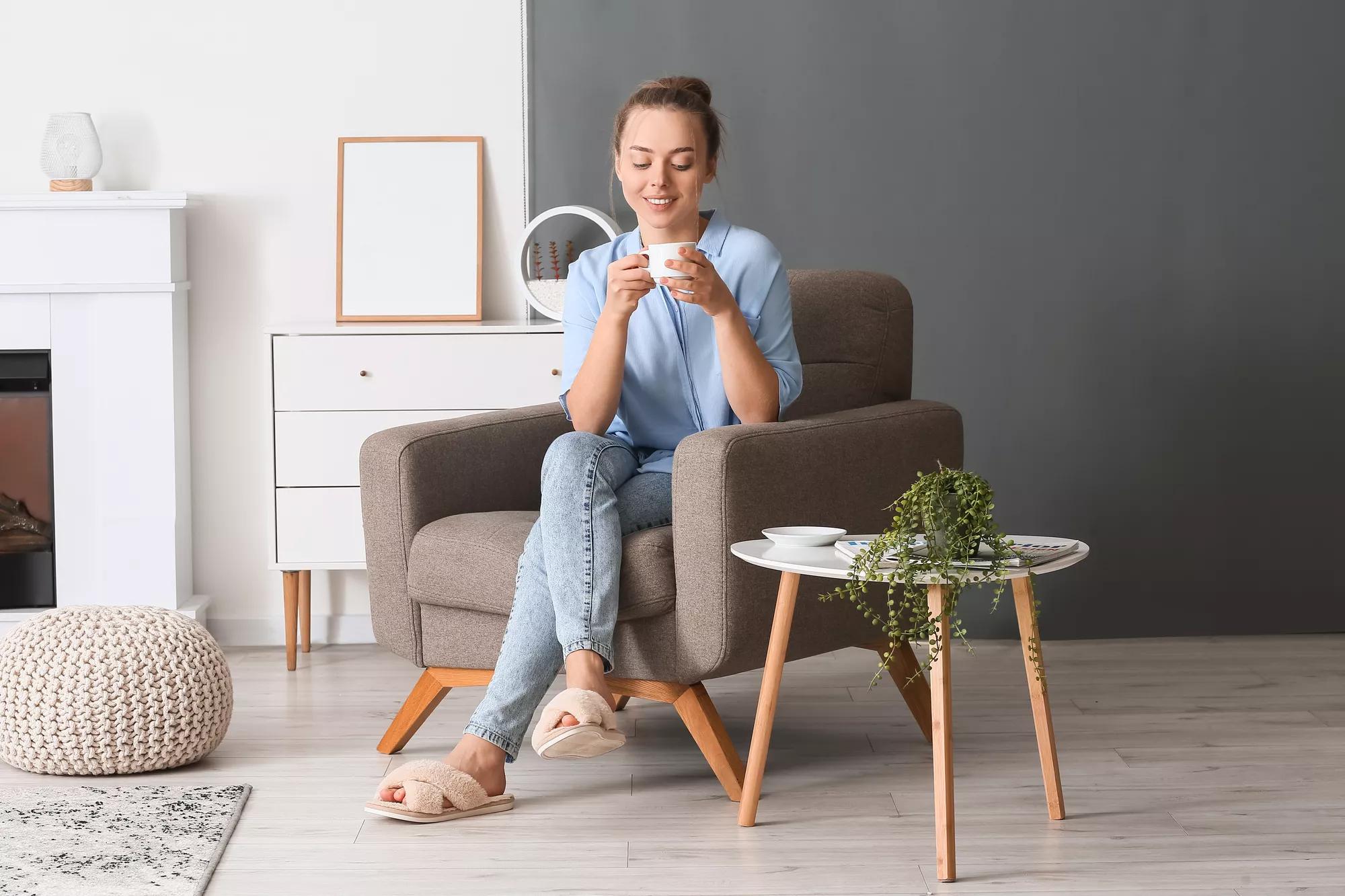 Young woman with cup of tasty coffee sitting in armchair at home