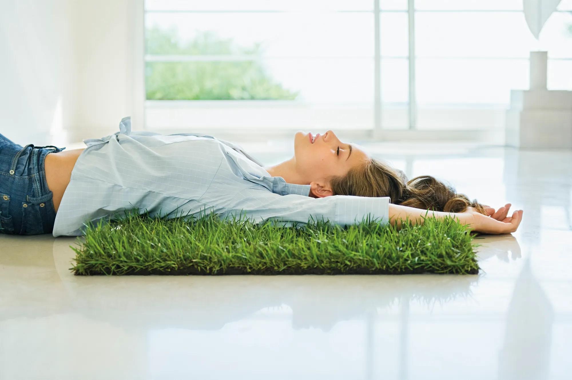 Profile of a girl 15 years old with long dark hair displayed and jeans laying down eyes closed and lips opened on a square made of grass placed on white ground, her light blue shirt is tied and raised up to show her belly, luminous interior and bay window in background, focus on girl, France, Nice