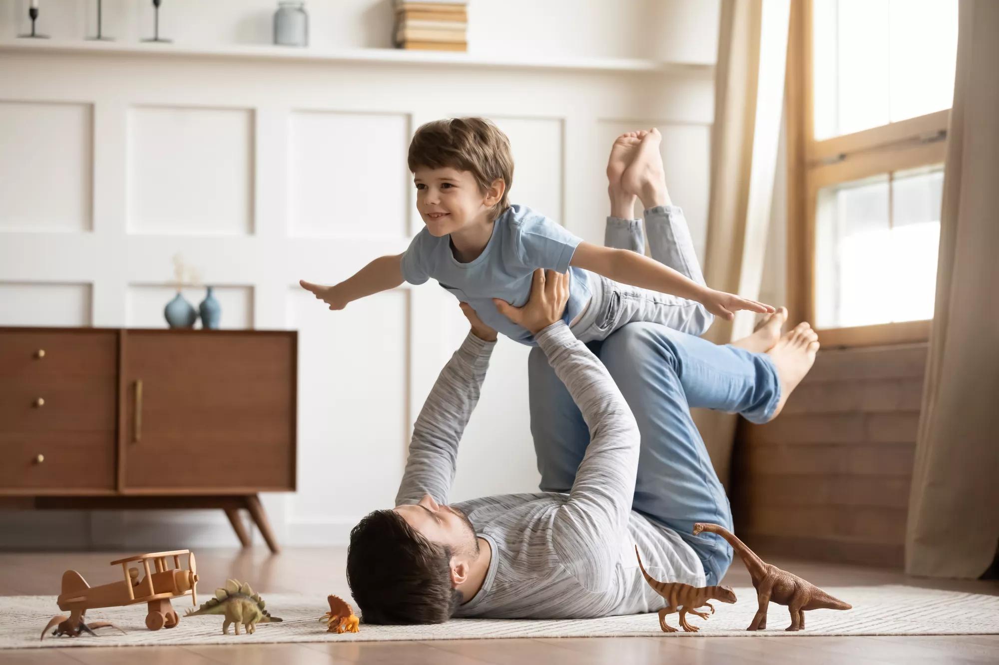 Joyful young man father lying on carpet floor, lifting excited happy little child son at home. Full length carefree two generations family having fun, practicing acroyoga in pair in living room.