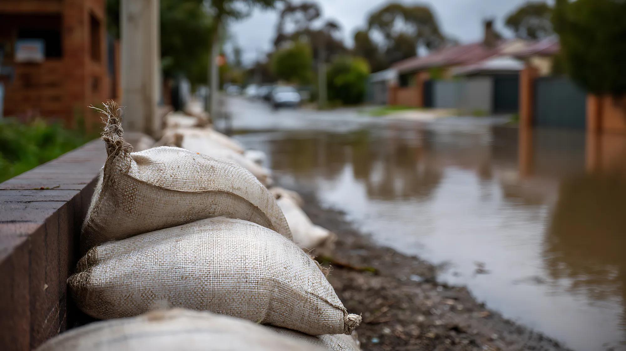 Close-up of stacked sandbags against a brick wall, acting as a barrier protecting properties from a flooded residential street. The murky water reflects houses under a cloudy sky, depicting a somber, protective, and challenging mood. Ideal for themes of natural disaster, climate change, community re