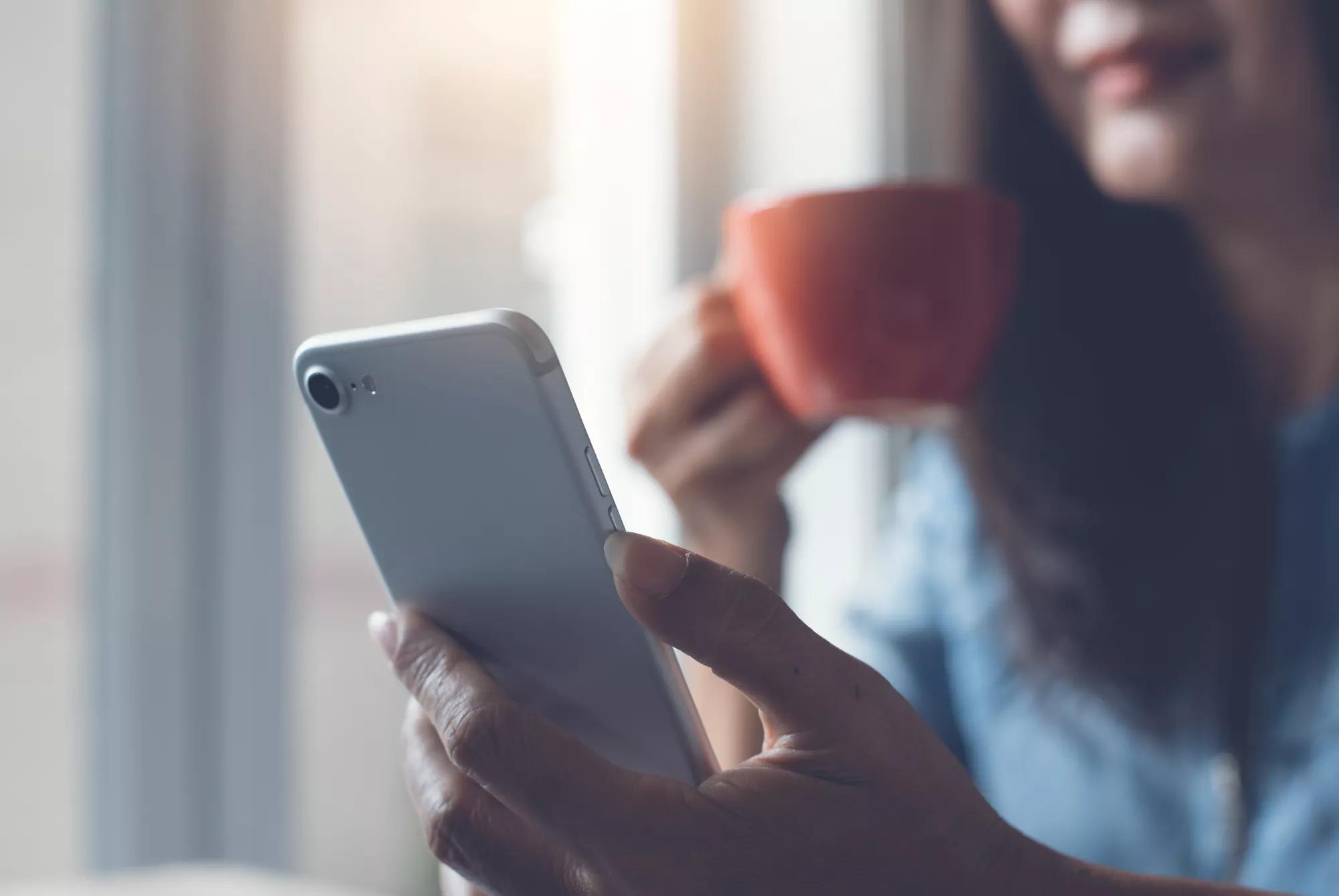 Cropped, business woman, entrepreneur holding red cup of hot coffee, morning news update via smart phone application at home, social media communication concept