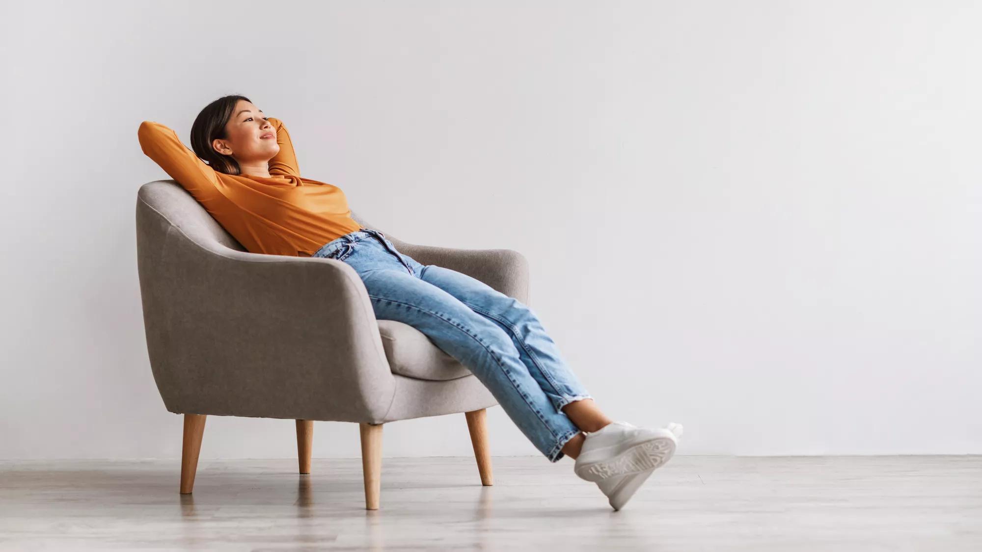 Gorgeous Asian woman sitting in comfy armchair, being lazy, feeling peaceful against white studio wall, panorama with copy space. Attractive young lady resting and relaxing with hands behind head