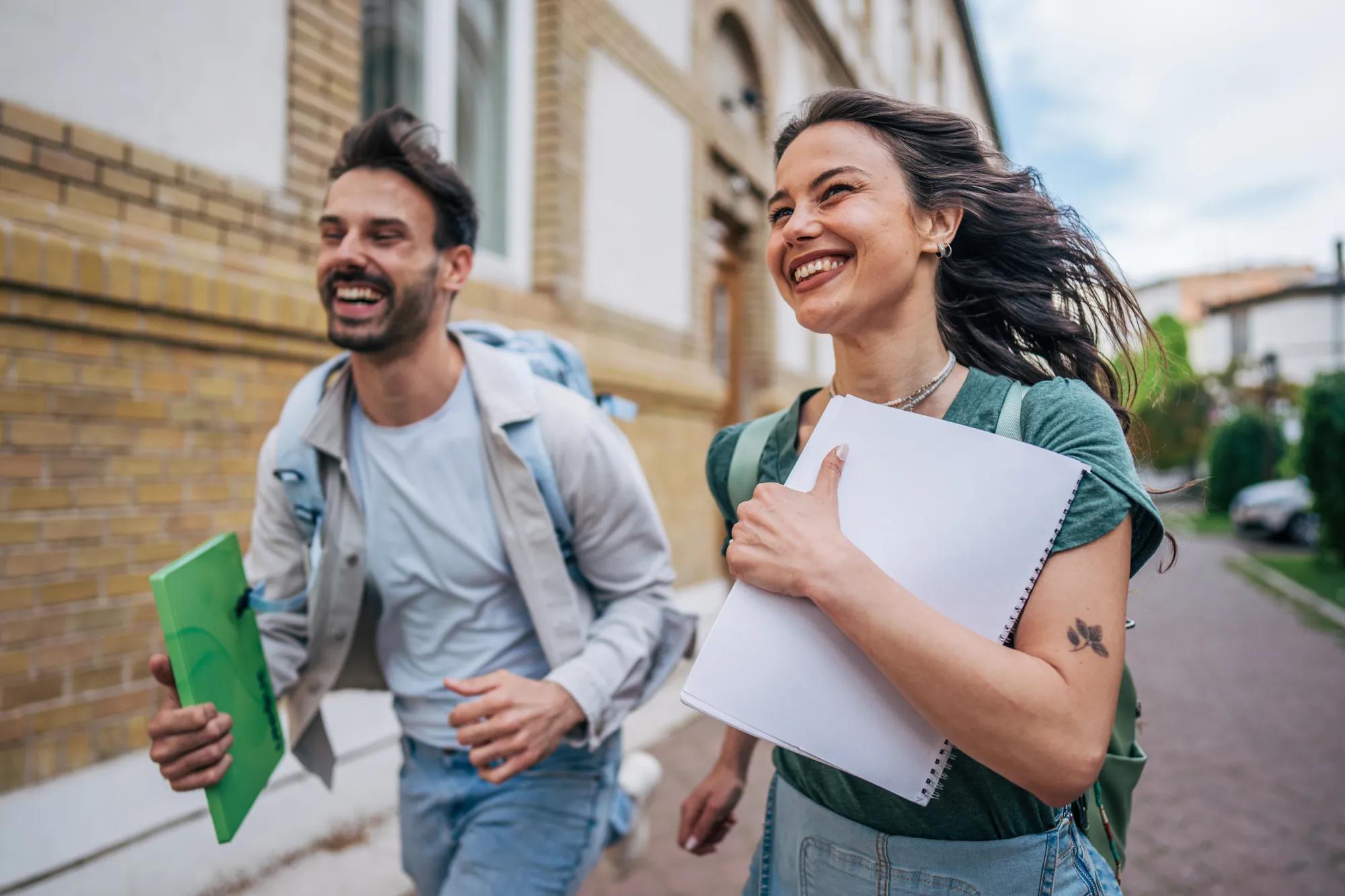 Two cheerful students running across a vibrant college campus, holding notebooks and backpacks, embodying the joy and freedom of university life while sharing laughter and friendship