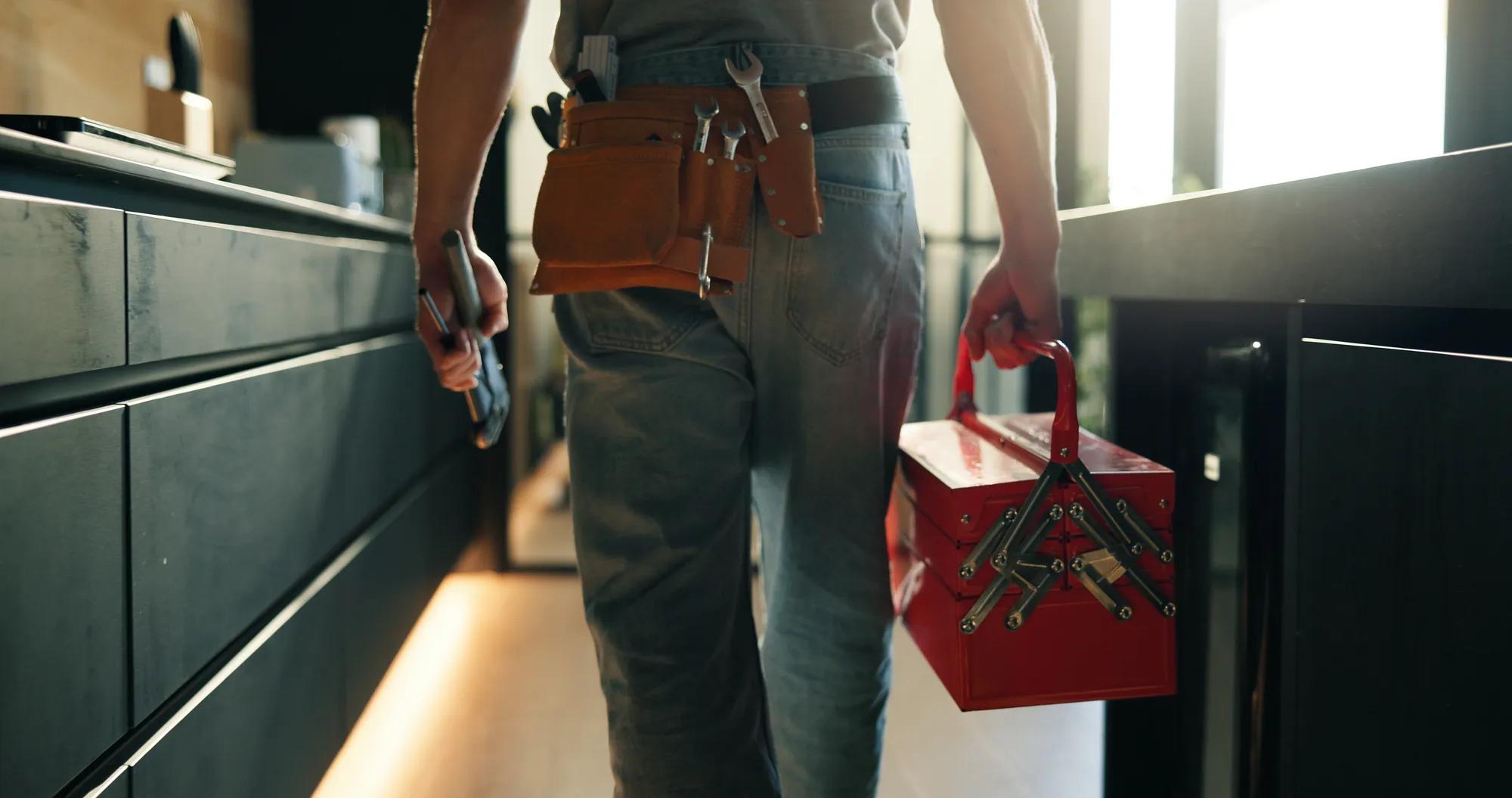 Walking, tool box and back of man in home for maintenance, service or repair job in kitchen. Equipment, customer care and male contractor with plumbing career at client house for leak inspection. AdobeStock_1841039889