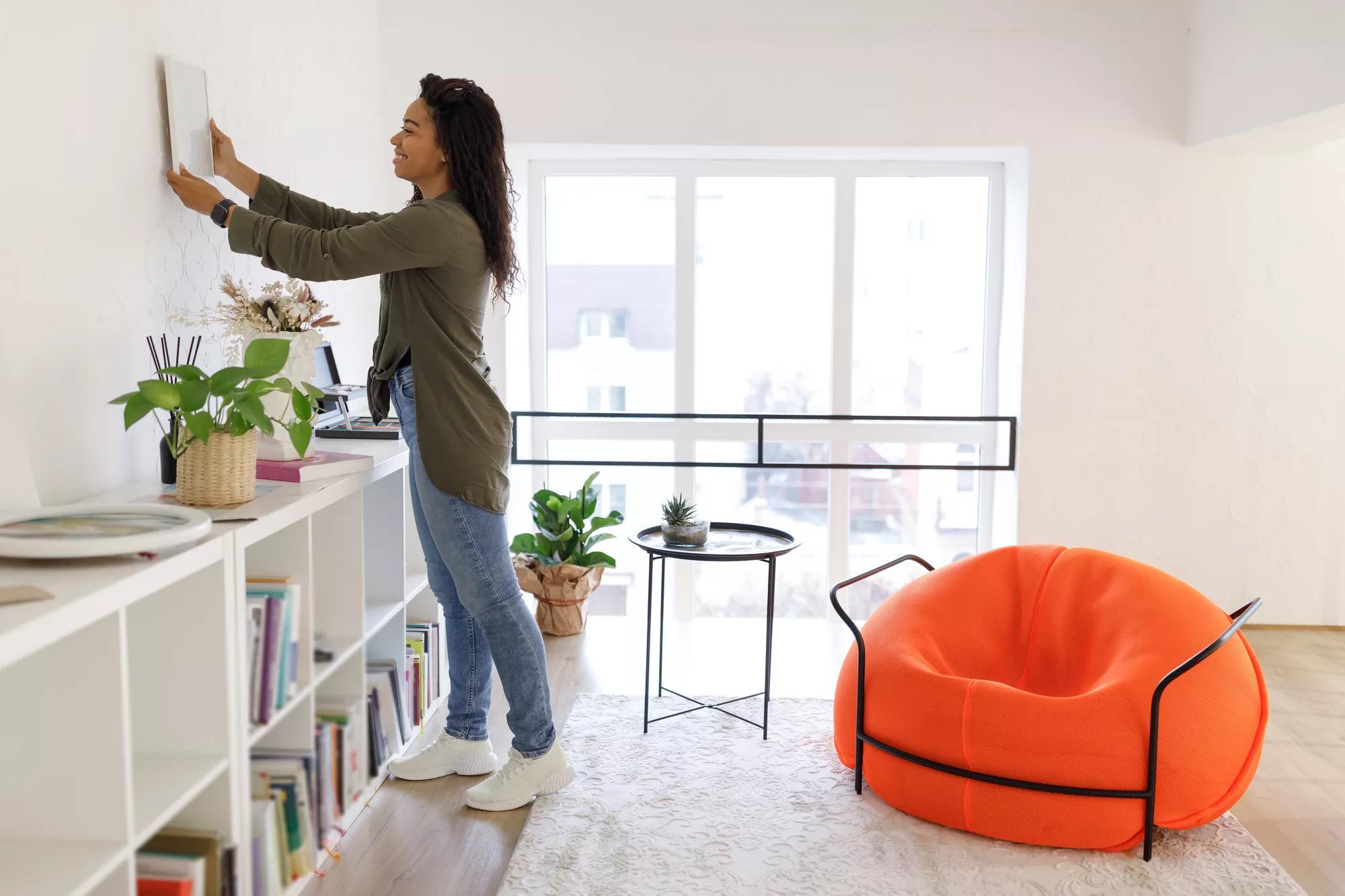 Home Interior And Domestic Decor. Smiling young black woman hanging painting, putting photo picture frame on the wall. Casual interior designer taking care of coziness in new modern stylish apartment