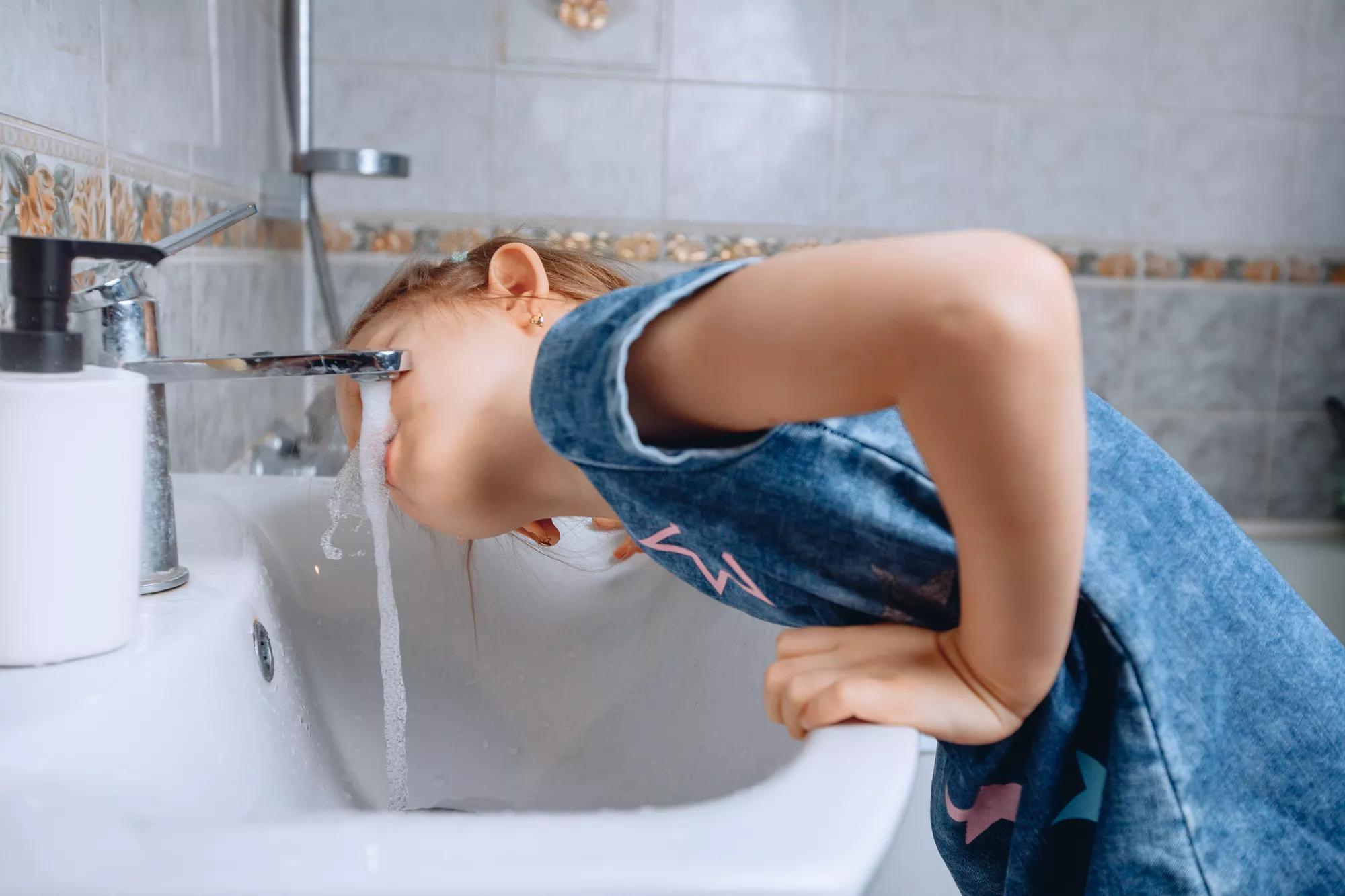 A little girl in a denim dress with a print leaned on a white ceramic sink to drink water from the tap in the bathroom.